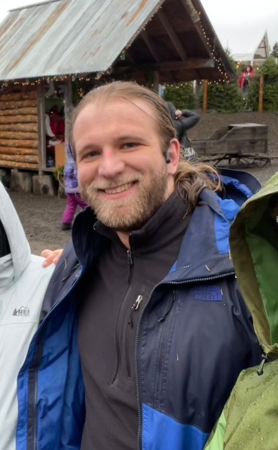 A man with long brown hair and a short-shorn beard. He is standing with others and smiling